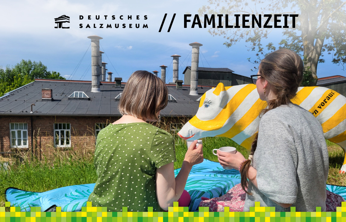 Picknick auf dem Solehügel, zwei Menschen sitzen mit Kaffeetassen auf einer Picknickdecke und blicken auf das historische Salinengebäude. Neben ihnen steht eine gelb-weiß-gestreifte Salzsau.